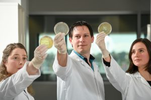 Sam Forster and Gemma D'Adamo and Tamblyn Thomason conducting UTI research holding up petri dishes at Hudson Institute.